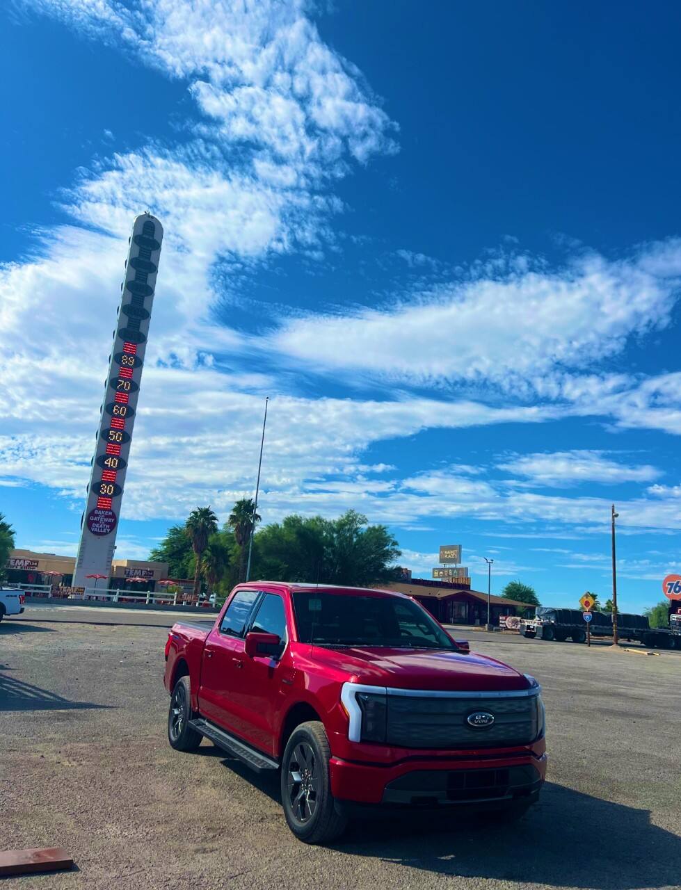 A red F-150 Lightning is parked near the World's Tallest Thermometer, which reaches into a blue sky with thin clouds, in Baker, California.